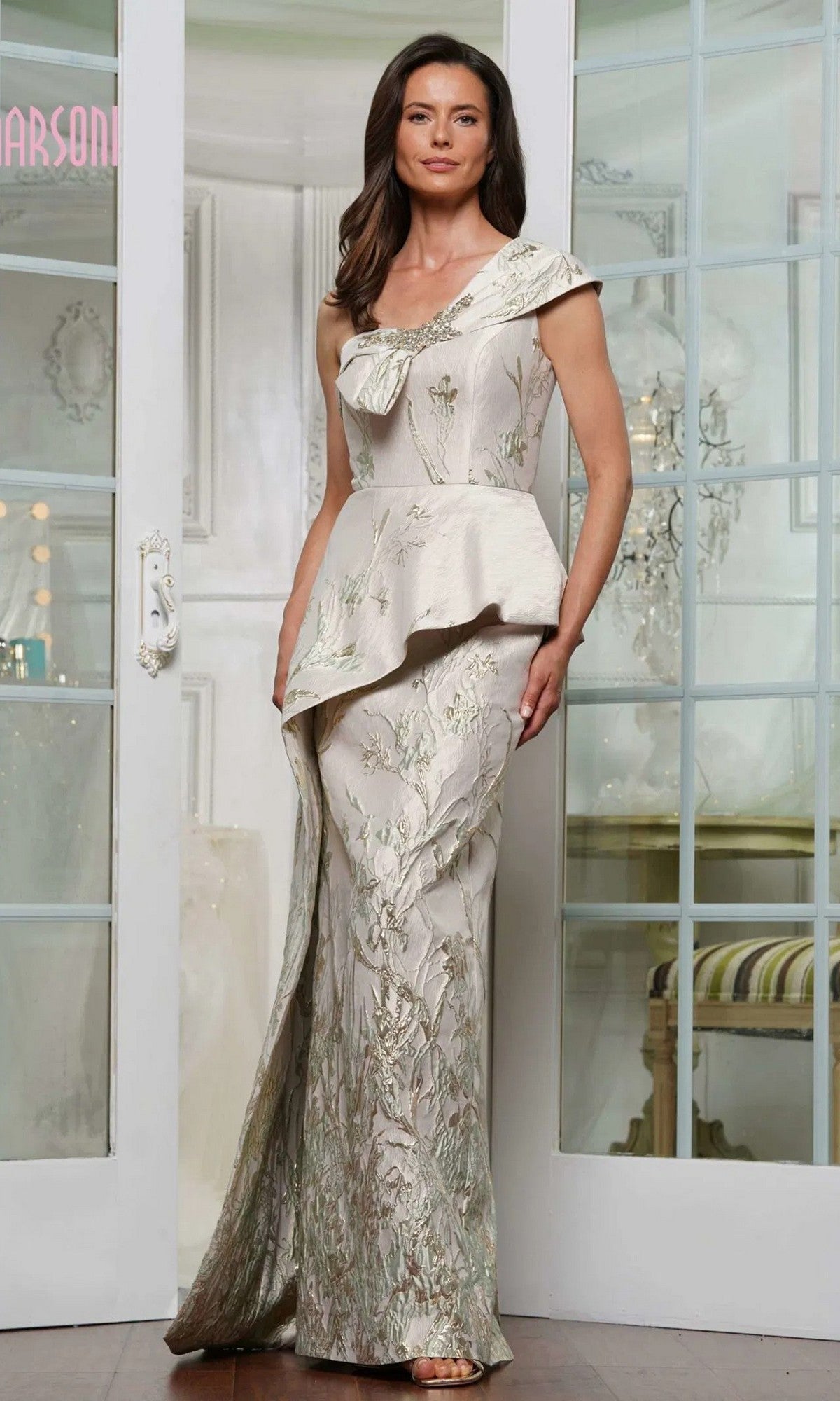 A woman stands indoors by glass doors wearing the Marsoni MV1300, a green gold long formal dress with floral embroidery, jacquard fabric, asymmetrical neckline, and peplum waist.